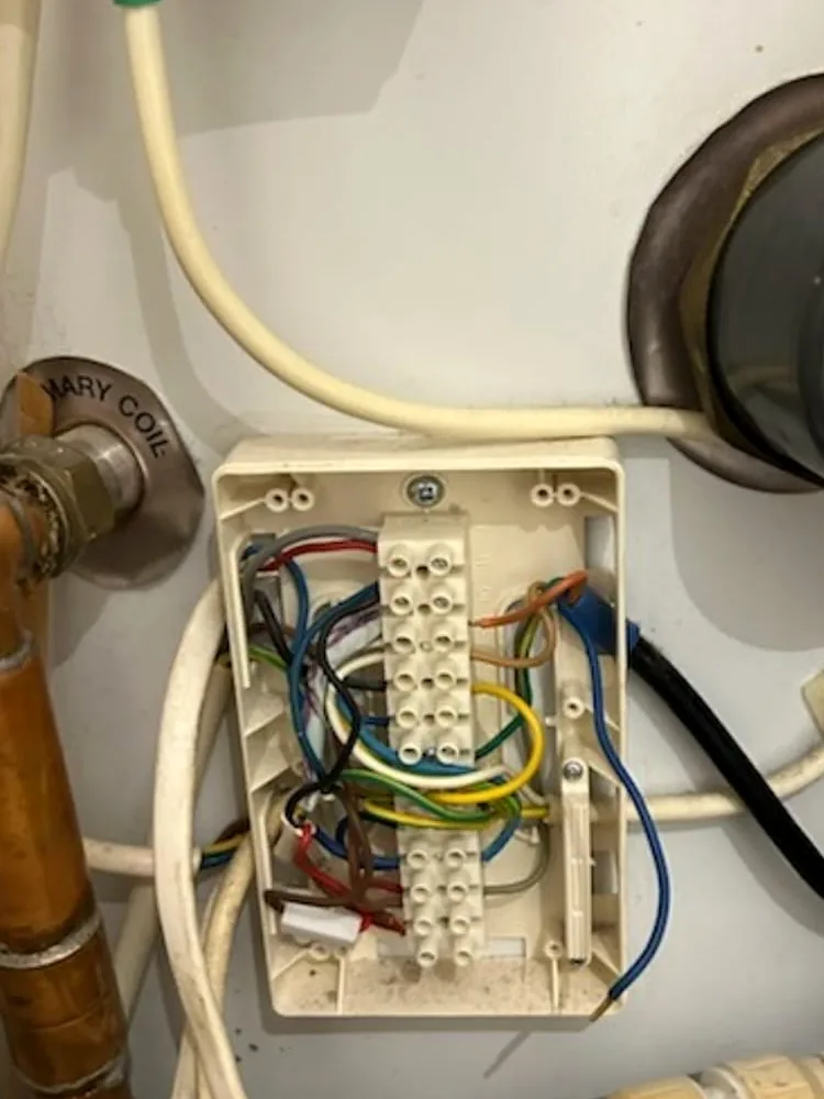 An open cream junction box on a white hot water cylinder, its terminal strips holding brown, blue, green-and-yellow, grey and red wires, with a white cable running down into it and the side of the immersion heater cap on the right.