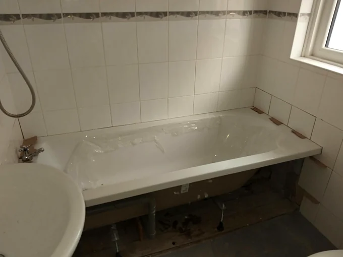 A new white bath fitted against a tiled wall, still in its protective film, with no front panel yet so its feet and the pipework underneath show, and pillar taps at the far end