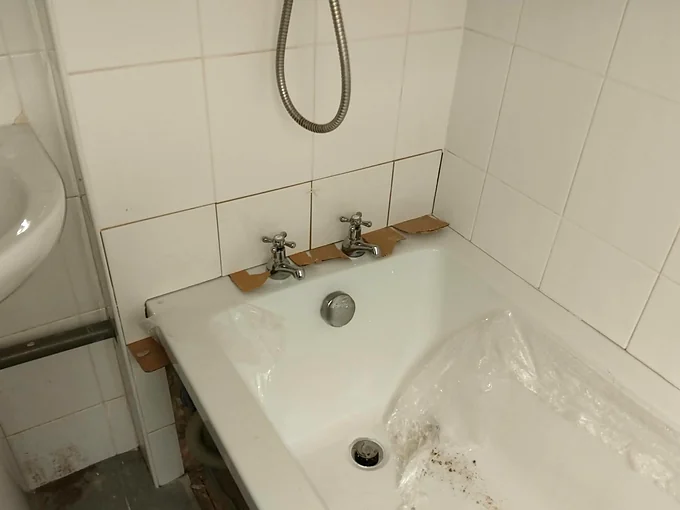 The tap end of the same bath, with two chrome cross head pillar taps, the plug and overflow fitted, and strips of cardboard along the joint between the bath and the tiles