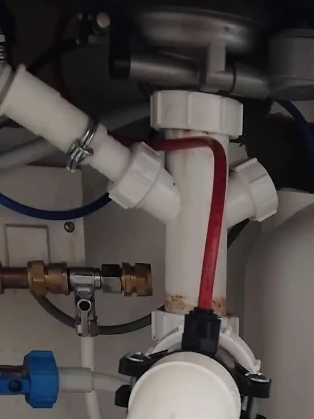 Under a kitchen sink: the bowl's metal underside and waste outlet, a white plastic waste pipe dropping through a junction, a red tube into a black clamp, a brass tee, a blue-handled valve and the white side of a water filter unit.