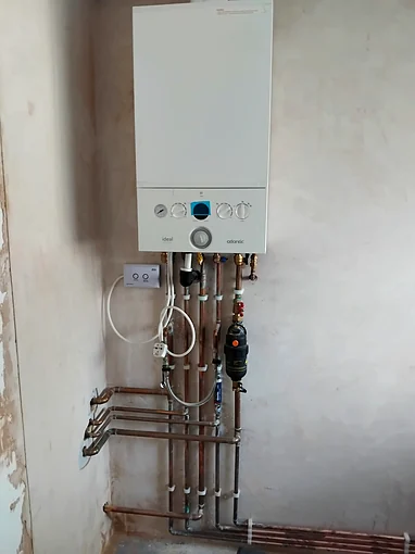 A white Ideal Atlantic combi boiler wall mounted on bare plaster, with copper pipework clipped in parallel runs beneath it, brass isolating valves, a black system filter and a white condensate pipe, and a wireless receiver and fused spur mounted to the left