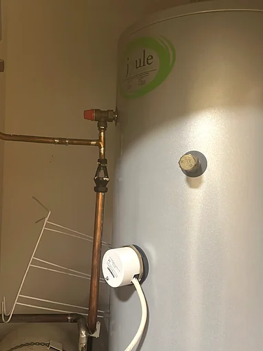 A grey Joule hot water cylinder in a cupboard, with the immersion element under a white cover near the bottom and its white cable, and copper pipework and a red capped relief valve to the left