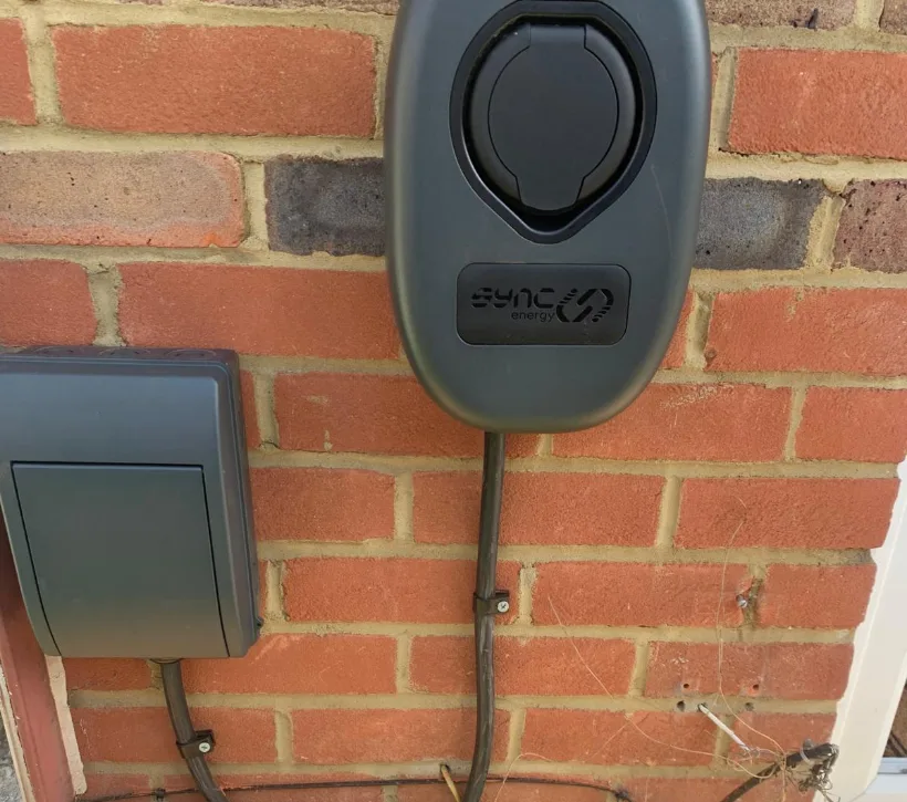 A black Sync Energy untethered EV charge point on a red brick wall, with a grey external isolator box beside it and armoured cable clipped down to ground level
