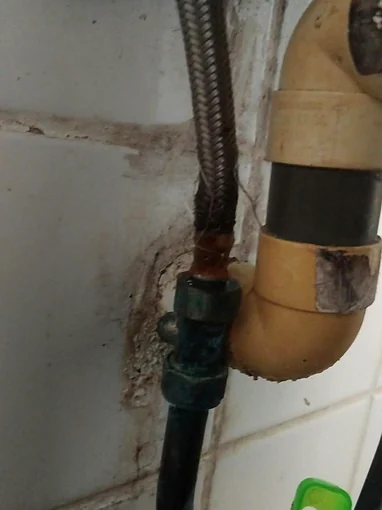 A braided steel flexi pipe under a sink, rusted brown and frayed at its lower end where it meets a valve crusted green, with a plastic waste trap beside it and stained tiles behind