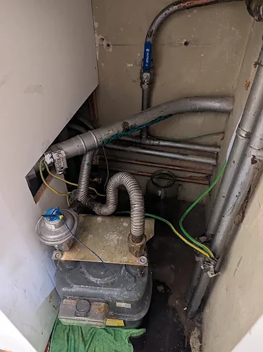 A gas meter in a cupboard with its regulator on the incoming pipe, two flexible connections on top of the meter, green and yellow earth bonding cables clamped to the pipework and grey pipes running up the wall