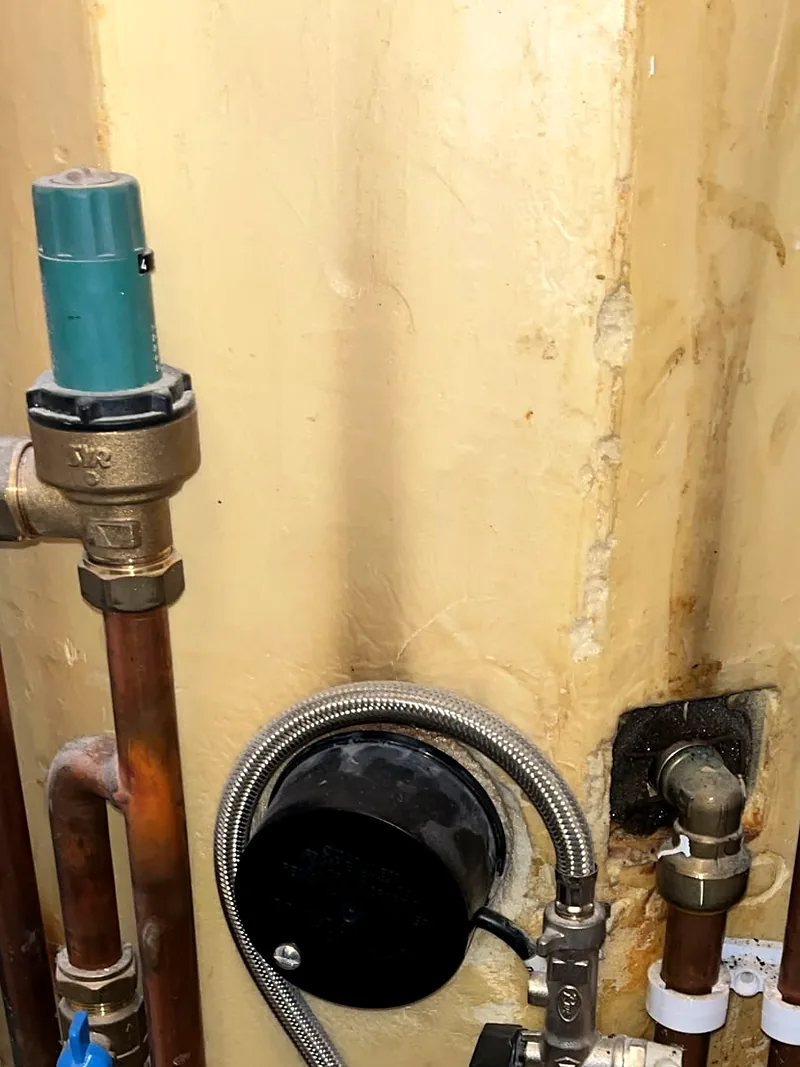 Lower part of a hot water cylinder in yellow foam insulation: copper pipes, a brass valve under a green cap, a braided flexible hose round a black cover, and rust stains on the foam round a brass pipe fitting.