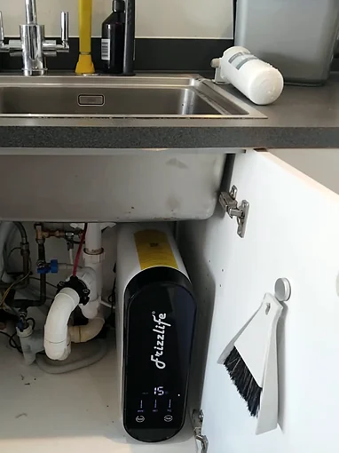 A white and black Frizzlife reverse osmosis unit standing upright in an open kitchen sink cupboard beside the waste pipework, with a slim black tap next to the chrome mixer on the sink above