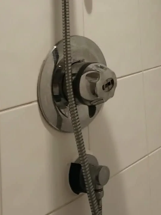 Close view of a round chrome shower mixer valve with a faceted turning knob on white tiles. The flexible metal shower hose runs past it, and a wall outlet elbow is fitted just below.