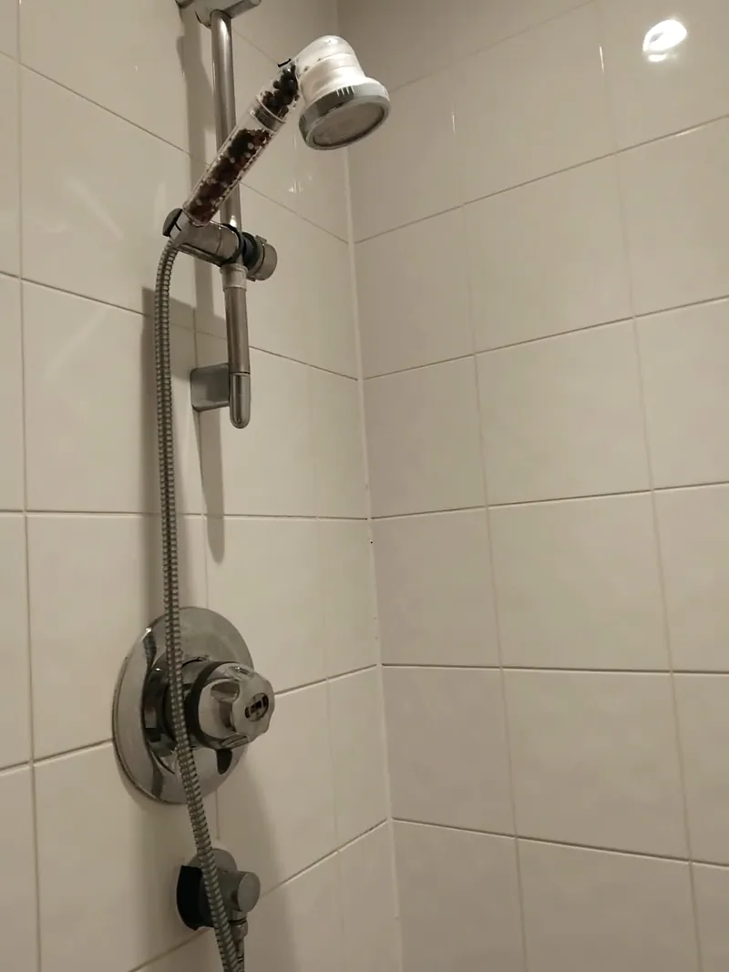 Shower corner with white tiles. A chrome riser rail holds a handheld shower head with a clear handle filled with beads. A metal hose runs down past a round chrome mixer valve to a wall outlet elbow.