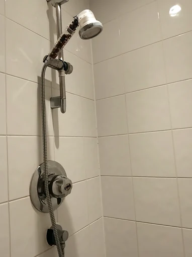 The same tiled corner before the work, with an old round chrome shower valve, a riser rail, a handset with a clear handle full of beads, and the hose looped down to an outlet on a black bracket