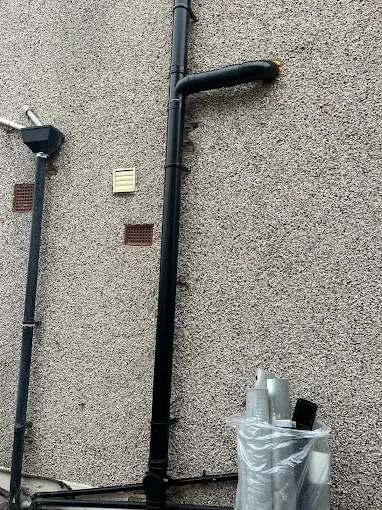 A black soil pipe running down a pebbledashed exterior wall with a swept bend at the top and cast brackets fixed into the render
