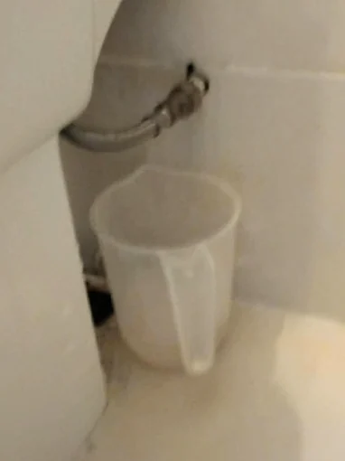Behind a toilet pan, a braided flexible feed pipe coming off the wall, and a plastic measuring jug on the floor underneath it
