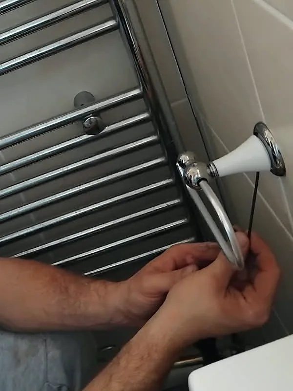 Close up of two hands holding the chrome arm of a toilet roll holder while a small hex key reaches up under its white post, the chrome wall plate on cream tiles and a chrome towel rail to the left