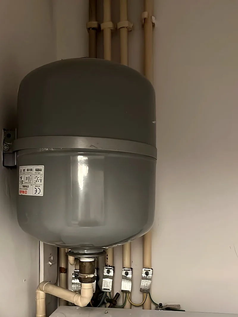 Grey expansion vessel held to a cupboard wall by a metal strap bracket. Its base has a brass fitting joined to cream pipework, and cream pipes with earth bonding clamps run up the wall behind it.