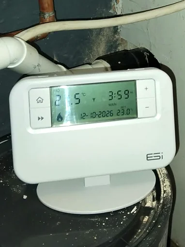A white ESi wireless programmable room thermostat on a desk stand, its display showing the temperature, the time, the date and a small flame symbol