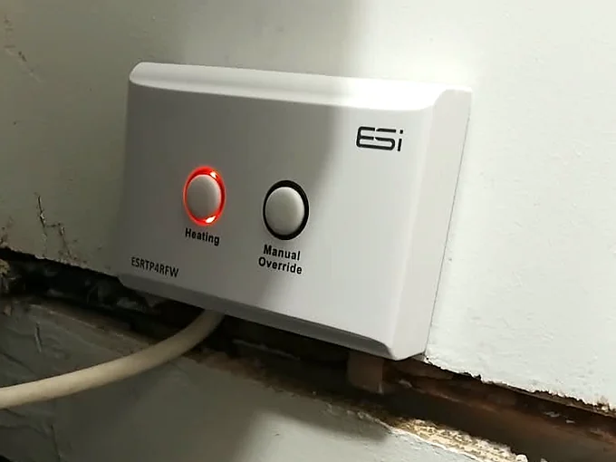 A white ESi receiver on a wall, the ring round its Heating button lit red, a Manual Override button beside it and a white cable running down from it
